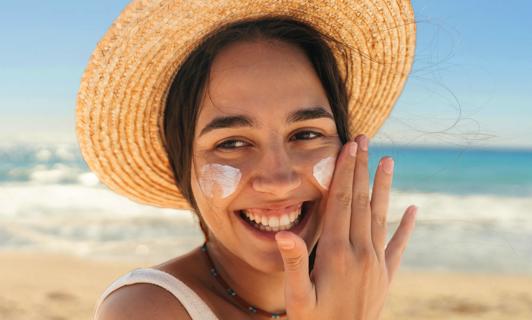 Smiling person wearing a straw hat at the beach, applying sunscreen to their face