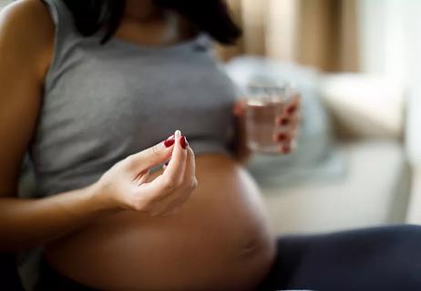 Pregnant woman holding a glass of water in one hand and a pill in the other.