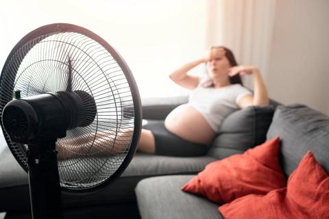 Fan blowing on pregnant person reclining on couch, with hand on forehead