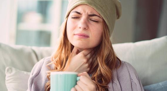 Person wincing as they hold their sore throat and a hot beverage