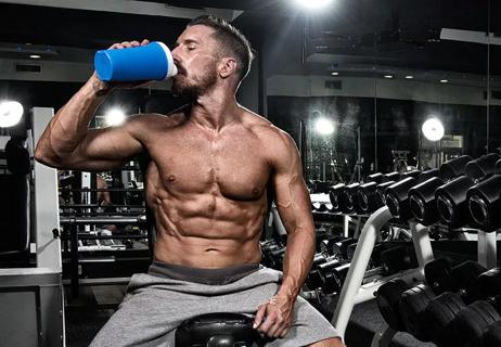 Bodybuilder drinks protein shake while sitting on weight bench with various hand weights in the background.