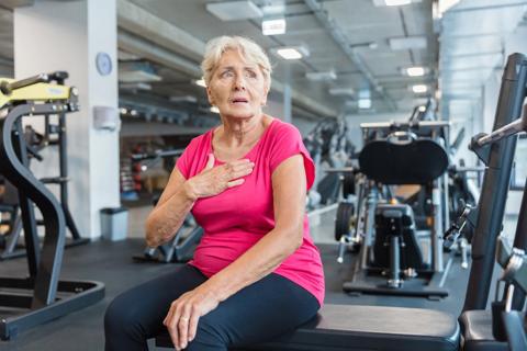 Older person sitting on exercise bench at gym, with hand on their chest