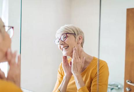 A smiling woman with short gray hair touching her cheeks while looking in the mirror