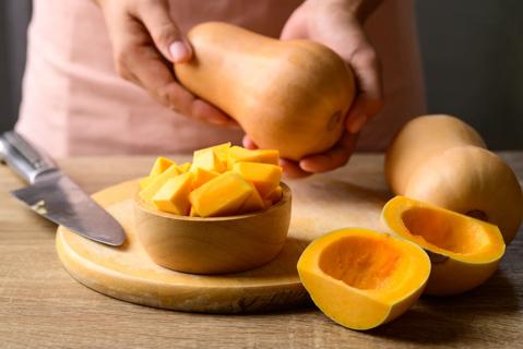 Person holding whole butternut squash, with bowl of cubed squash and sliced squash on table