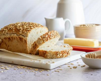 Partially sliced loaf of oats and honey bread on cutting board, with honey, butter nearby, oats sprinkled on table