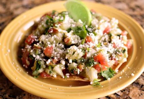 Fish tostada on a yellow plate piled with tomatoes, avocados and a lime.