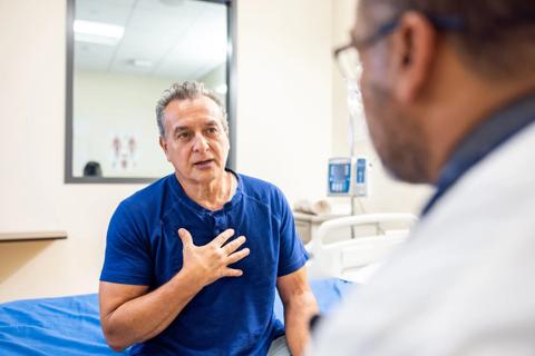 Patient in exam room with hand on chest, talking to healthcare provider