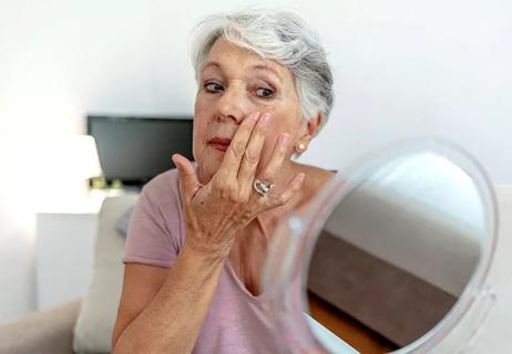 Senior woman looks in a small mirror while examing the skin beneath her eyes