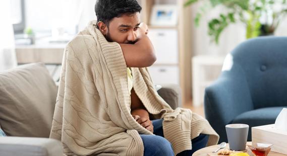 Person sitting on couch, wrapped in blanket, coughing into elbow, with tissues and hot beverage nearby