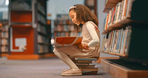 Child sitting on stack of books reading a book in the library