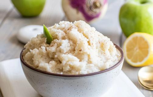 A bowl of cooked mashed turnips and Granny Smith apples, with lemons, apples and turnips in background