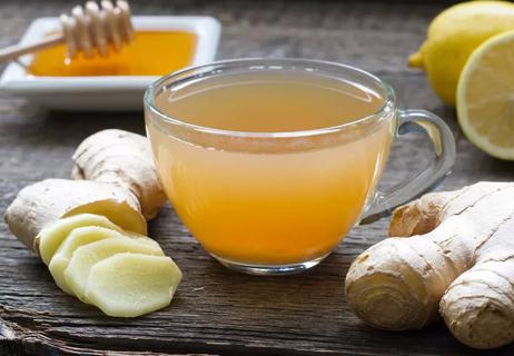 Ginger tea in a clear glass cup surrounded by ginger root with honey and lemons in the background.