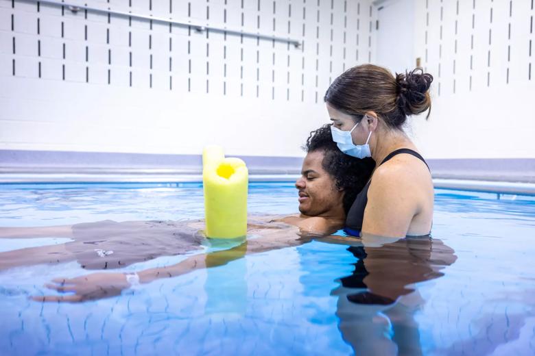 Person helps another person float in the pool with a yellow foam noodle