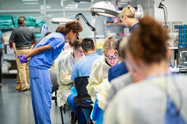 A surgeon bends down to instruct students practicing surgical techniques