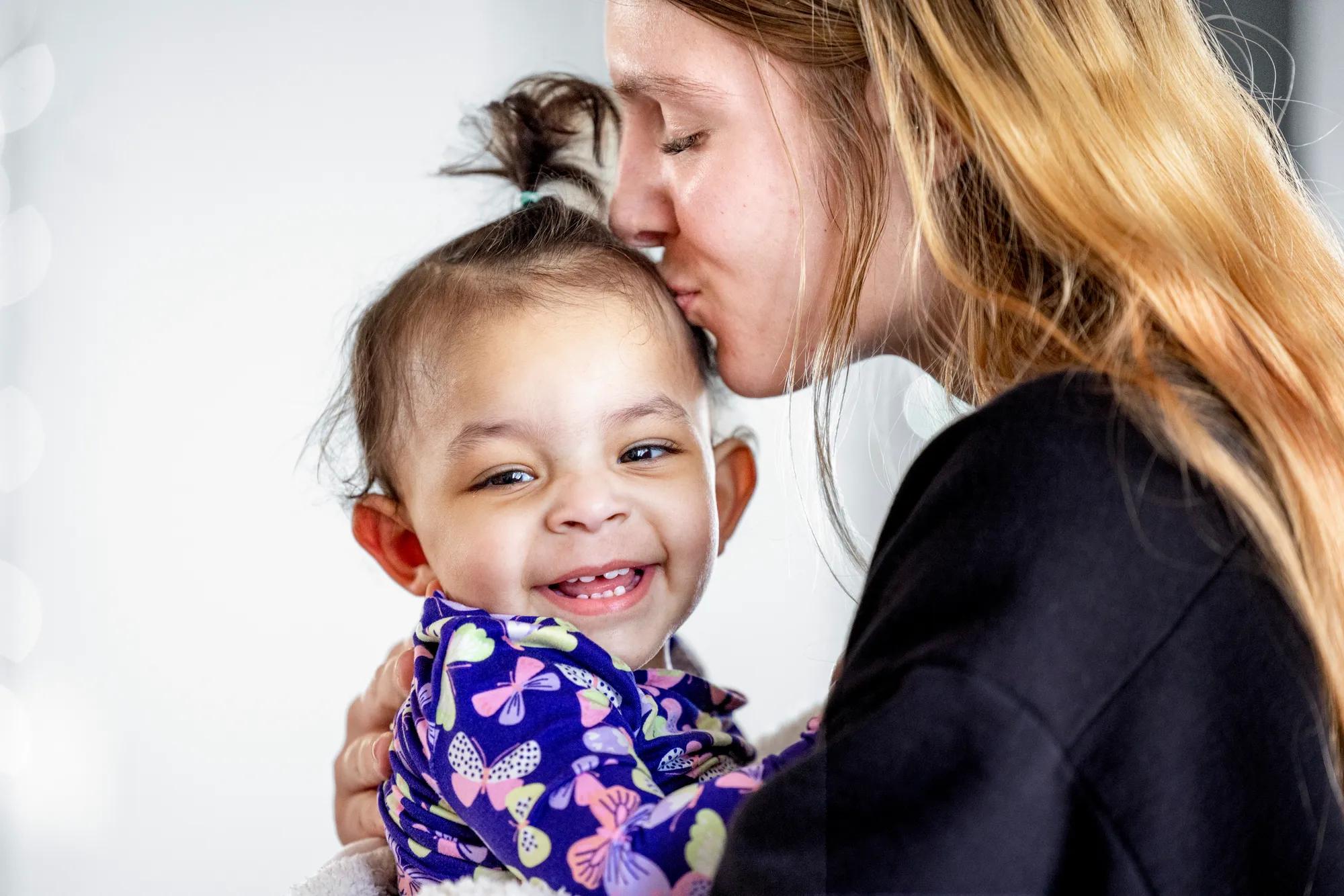 A mother kisses her smiling toddler wearing a purple butterfly shirt.