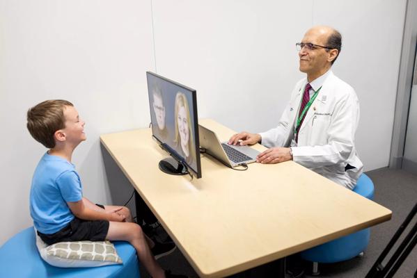 A man in a white lab coat sitting across a desk from a child staring at a screen.