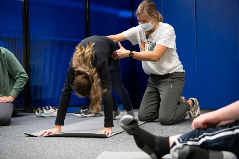 Woman kneeling on the floor pulls up the torso of someone bending over with both feet and both hands on the floor