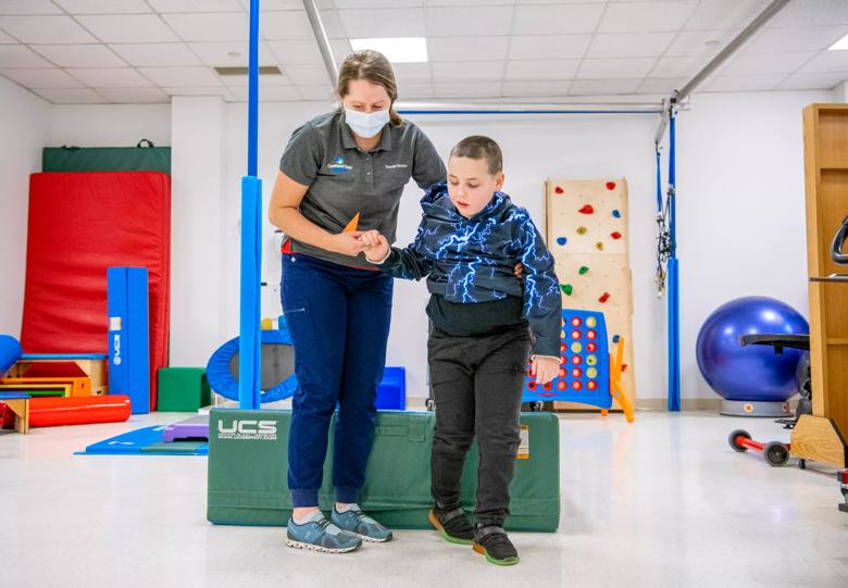 A caregiver helps a child walk in a room with therapy toys