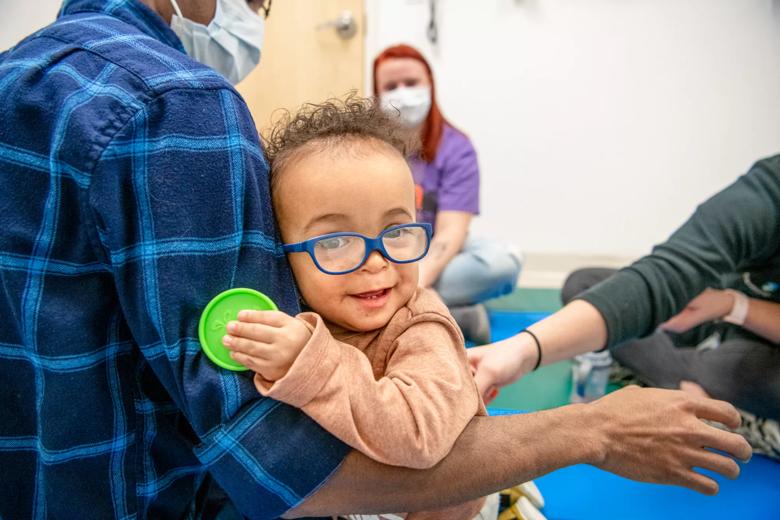 Little boy in blue glasses holds a green disk while hugging an adult