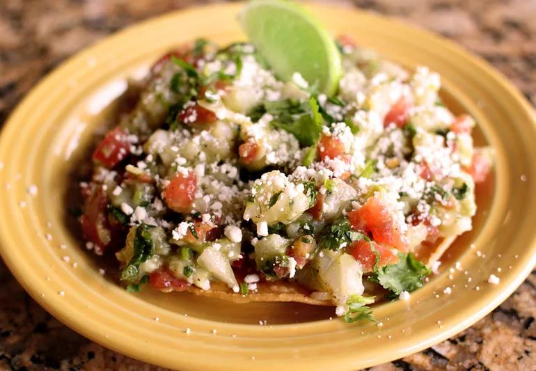 Fish tostada on a yellow plate piled with tomatoes, avocados and a lime