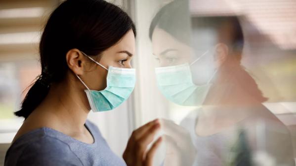 Portrait of sad young woman with face protective mask looking through the window at home